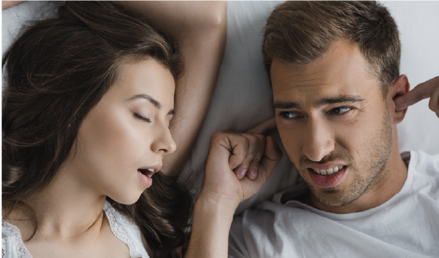 A young woman snoring next to a man who is sticking his fingers in his ears